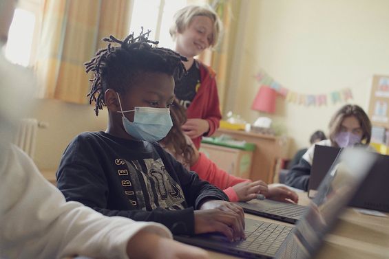 Kind mit Maske am Laptop in einem Raum mit anderen Kindern
